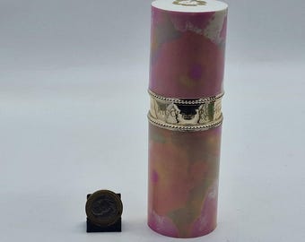 May include: A cylindrical perfume bottle with a pink and gold floral design. The bottle has a gold band with the text "ARTCO Paris" and a white top. A small coin is next to the bottle.