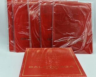 May include: A collection of red boxes and packaging, some wrapped in clear plastic. The boxes have gold lettering that reads "BALENCIAGA PARIS". The items are arranged on a white surface.