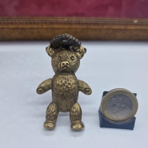Op de afbeelding: Een kleine, antieke messing teddybeerfiguur met een getextureerd oppervlak. De beer heeft een kleine hoed en staat rechtop. Een euro munt ligt naast de beer ter vergelijking van de grootte. De beer is ongeveer 5 cm hoog.