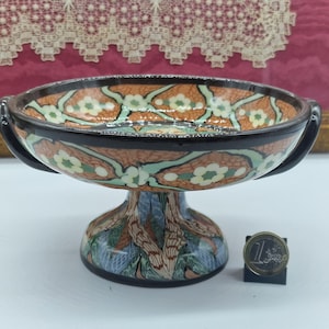 May include: A decorative ceramic pedestal bowl with a detailed floral pattern in orange, green, and white against a brown background. The bowl has a black rim and handles, and a patterned base. A coin is next to the bowl.