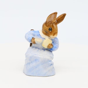 Puede incluir: Una figurita de conejo marrón con un vestido azul y sosteniendo una taza y un libro. La figurita está sentada en una silla.