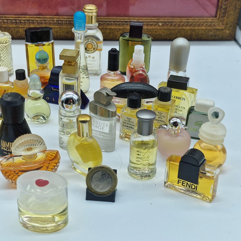 K&ouml;nnte beinhalten: Eine Sammlung von Miniatur-Parf&uuml;mflaschen in verschiedenen Formen und Gr&ouml;&szlig;en. Die Flaschen sind mit farbigen Fl&uuml;ssigkeiten gef&uuml;llt und weisen unterschiedliche Verschlussdesigns auf. Einige Flaschen haben goldene Akzente, und eine M&uuml;nze ist ebenfalls vorhanden.