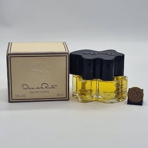 Peut inclure: Flacon de parfum Oscar de la Renta Eau de Toilette avec un bouchon noir et une boîte beige. Le flacon contient un liquide jaune et est étiqueté "60 ml". La boîte est beige avec le nom de la marque et un motif floral.