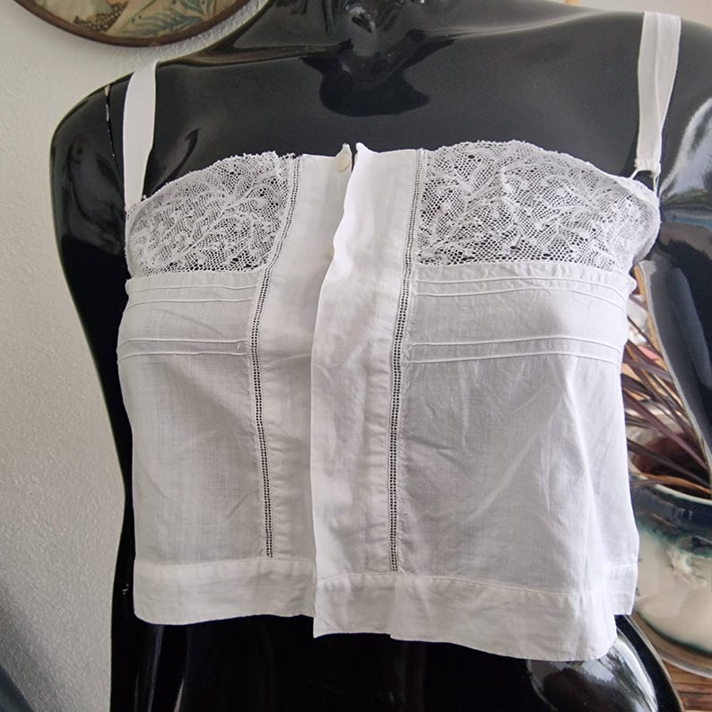 May include: White cotton bustier with lace detailing at the top. The bustier has thin, adjustable straps and a front closure. The garment features vertical seams and horizontal pleats, giving it a structured appearance. The bustier is displayed on a black mannequin.