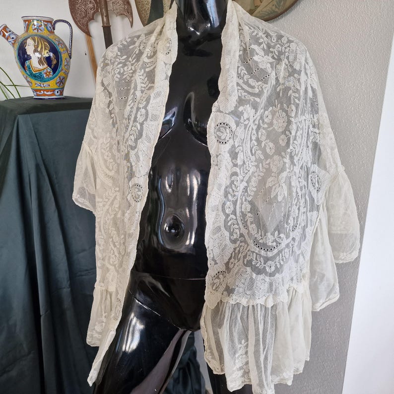 May include: Cream-coloured lace shawl with floral and geometric patterns. The shawl has short sleeves and a ruffled hem. The delicate fabric is draped over a black mannequin, showcasing the intricate design and texture of the vintage garment.