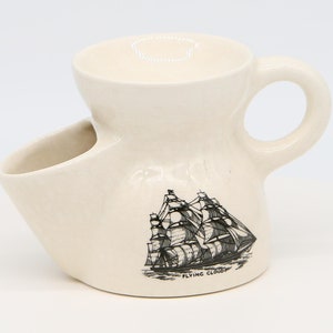 Peut inclure: Une tasse à raser en céramique blanche avec une poignée et un porte-savon intégré. La tasse présente une illustration en noir et blanc d'un voilier avec le texte "Flying Cloud" en dessous.