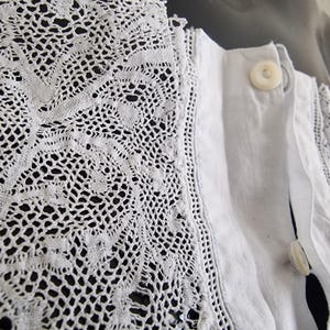May include: Close-up of a white garment featuring intricate lace detailing along the collar and edges. The lace has a delicate, openwork pattern. The garment has a button closure with two visible buttons.