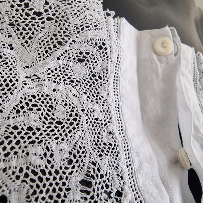 May include: Close-up of a white garment featuring intricate lace detailing along the collar and edges. The lace has a delicate, openwork pattern. The garment has a button closure with two visible buttons.