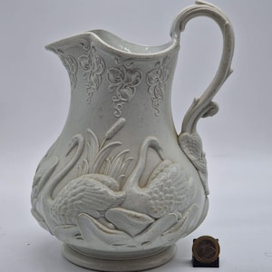 Può includere: Brocca in ceramica color crema con un design a rilievo dettagliato di cigni, giunchi e fogliame. La brocca ha un manico curvo e un bordo decorativo. La base è rotonda e la brocca è alta circa 25 cm.