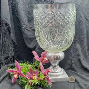 Op de afbeelding: Een grote, heldere kristallen vaas met ingewikkelde diamant- en waaiervormige snijpatronen. De vaas staat op een zilverkleurige voet. Een boeket roze orchideeën en groen is voor de vaas geplaatst.