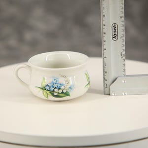 Puede incluir: Una taza de cerámica blanca con un patrón floral azul y verde de lirios de los valles. Una regla de metal está colocada junto a la taza, indicando una altura de aproximadamente 6,5 cm para el recipiente, que tiene una sola asa.