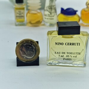K&ouml;nnte beinhalten: Eine Sammlung von Miniatur-Parf&uuml;mflaschen, darunter eine quadratische Nino Cerruti Eau de Toilette-Flasche mit schwarzem Verschluss. Die Flasche enth&auml;lt eine hellgelbe Fl&uuml;ssigkeit und hat Text auf dem Etikett. Eine Euro-M&uuml;nze wird in einem schwarzen St&auml;nder gezeigt.