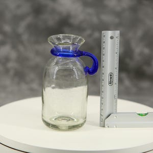 Peut inclure: Une carafe en verre transparent avec une anse en verre bleu et un col évasé. Une règle en métal argenté est placée à côté de la carafe, indiquant une hauteur d'environ 14 cm. La carafe a un corps arrondi et un col étroit.