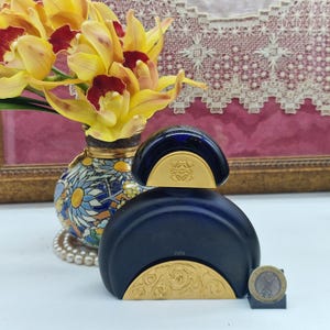 May include: A navy blue perfume bottle with gold detailing and a gold emblem. The bottle is next to a coin and a vase with yellow and red flowers.