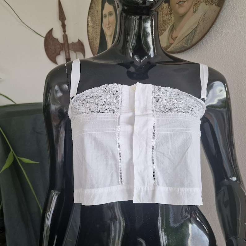 May include: A white cotton bustier with lace detailing across the top. The bustier has thin straps and a button-up front. The garment is displayed on a black mannequin. The bustier is a vintage style.