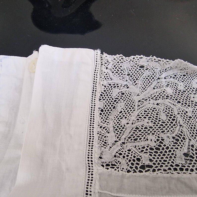 May include: Close-up of white fabric with intricate lace detailing. The lace is on the right side of the image, with a delicate pattern. The left side shows a folded section of plain white fabric with a button.