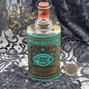 May include: A vintage glass bottle of Eau de Cologne, featuring a label with teal and gold accents. The label reads "4711" and includes text in German. The bottle has a red and gold cap. A coin is next to the bottle.