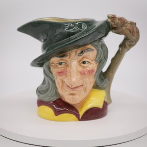 May include: A ceramic jug shaped like a woman's head wearing a green hat and a yellow and red shirt. The jug has a handle shaped like a branch.