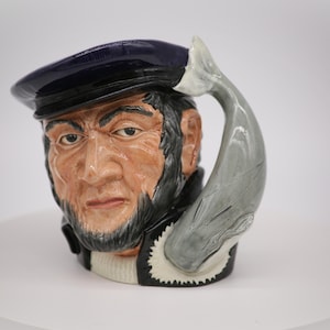 May include: A ceramic mug shaped like a man's head wearing a navy blue cap. The mug has a whale tail handle.