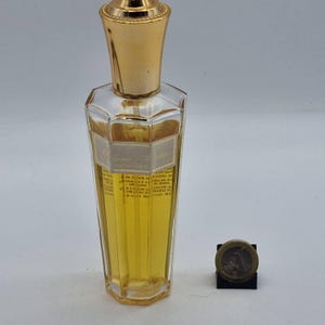 Könnte beinhalten: Eine klare, achteckige Parfümflasche mit goldfarbenem Verschluss. Die Flasche enthält eine gelbe Flüssigkeit und hat ein Etikett mit Text. Neben der Flasche liegt eine Münze.