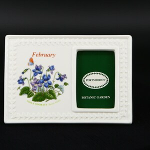 May include: White ceramic picture frame with a floral design and the word "February" printed on it. The frame has a small opening for a photo and a green background with the text "Portmeirion Botanic Garden".