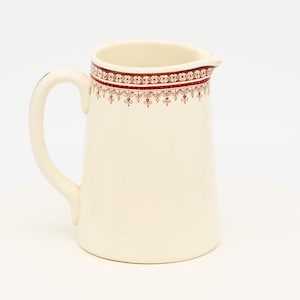 Op de afbeelding: Crèmekleurige keramische kan met een handvat en tuit. De bovenrand heeft een decoratieve band met rode en bruine bloemmotieven. Een klassiek stuk voor het serveren van dranken.