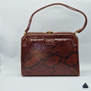 May include: A vintage brown snakeskin handbag with a rectangular shape and a top handle. The bag features a gold-coloured clasp and a small front pocket. The snakeskin pattern is a mix of brown and black.