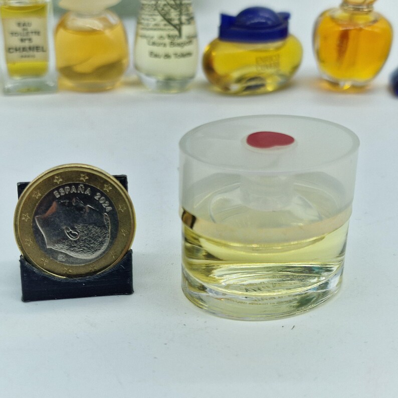 K&ouml;nnte beinhalten: Ein klares Parf&uuml;mfl&auml;schchen mit einem goldenen Band und einem roten Deckel, gef&uuml;llt mit einer gelben Fl&uuml;ssigkeit. Eine Euro-M&uuml;nze in einem schwarzen Halter befindet sich neben der Flasche. Weitere Miniatur-Parf&uuml;mflaschen sind im Hintergrund.