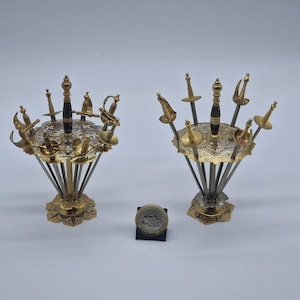 May include: Two decorative sword holders with miniature swords. The holders are gold-toned with intricate designs and a central sword with a black handle. A coin is placed in front of the holders for scale.