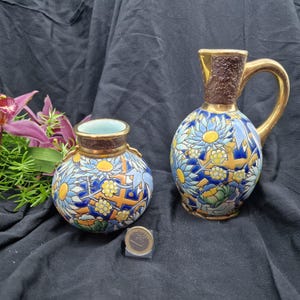 May include: Two antique ceramic vases with floral designs. One is a small, round vase, and the other is a pitcher with a handle. Both have blue, yellow, and orange floral patterns on a white background. The pitcher is approximately 20 cm tall.