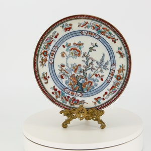 Puede incluir: Un plato de porcelana blanca con un diseño floral azul y rojo. El plato está sobre un soporte de metal dorado.