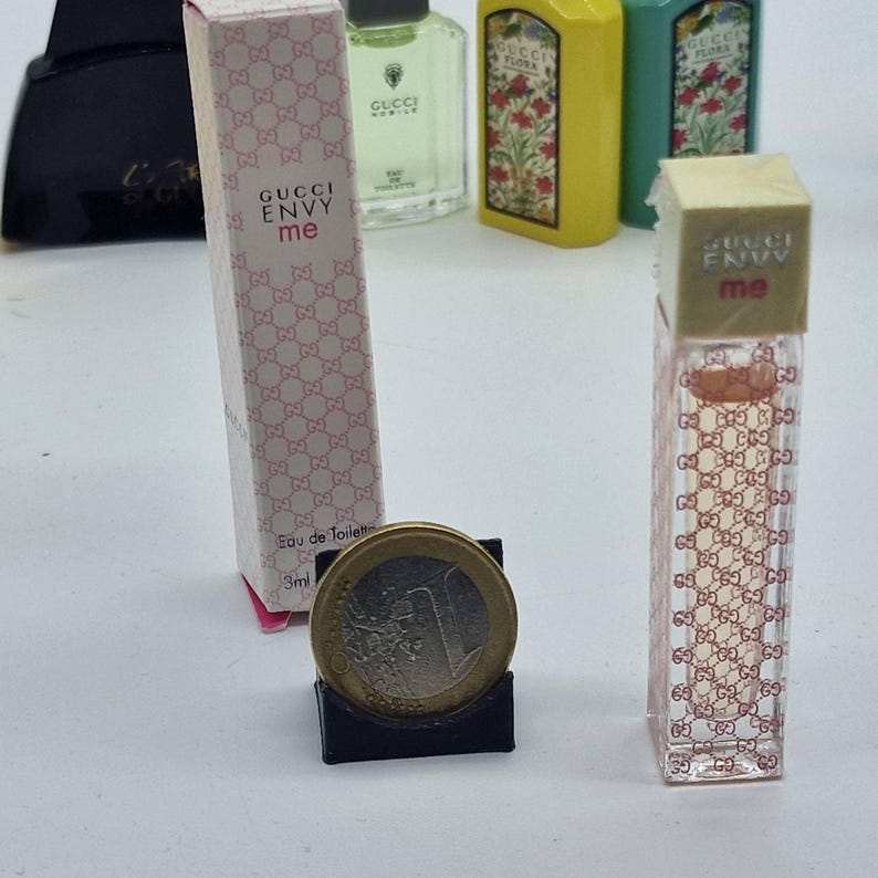 May include: A miniature perfume bottle and box of Gucci Envy Me Eau de Toilette. The box and bottle are pink and feature the Gucci logo. A coin is in the foreground. Other perfume bottles are in the background.
