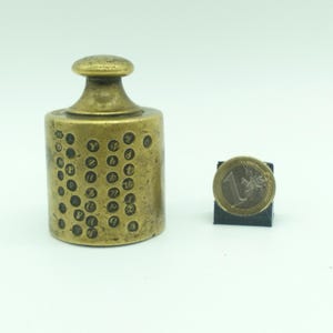 Op de afbeelding: Een klein, rond, messing gewicht met een knop aan de bovenkant. Het gewicht is bedekt met kleine, ronde inkepingen met daarin gegraveerde cijfers en letters.