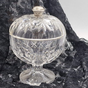 Peut inclure: Un bonbonnière en verre transparent avec un couvercle argenté et un motif en verre taillé. Le plat est sur un fond de dentelle noire.