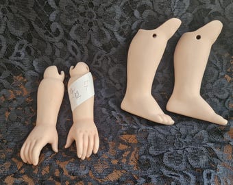 Op de afbeelding: Een verzameling vintage poppenledematen, waaronder twee benen en twee armen. De ledematen zijn in een bleke huidskleur, met gedetailleerde vingers en tenen. De armen hebben een papieren label met handgeschreven nummers. Op donkere, bedrukte stof.