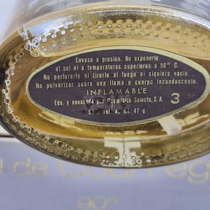 May include: Close-up of a perfume bottle with a gold-colored liquid. The bottle has a dark label with Spanish text, including warnings about pressure and flammability. The label also indicates a volume of 45 ml and a weight of 47 g.