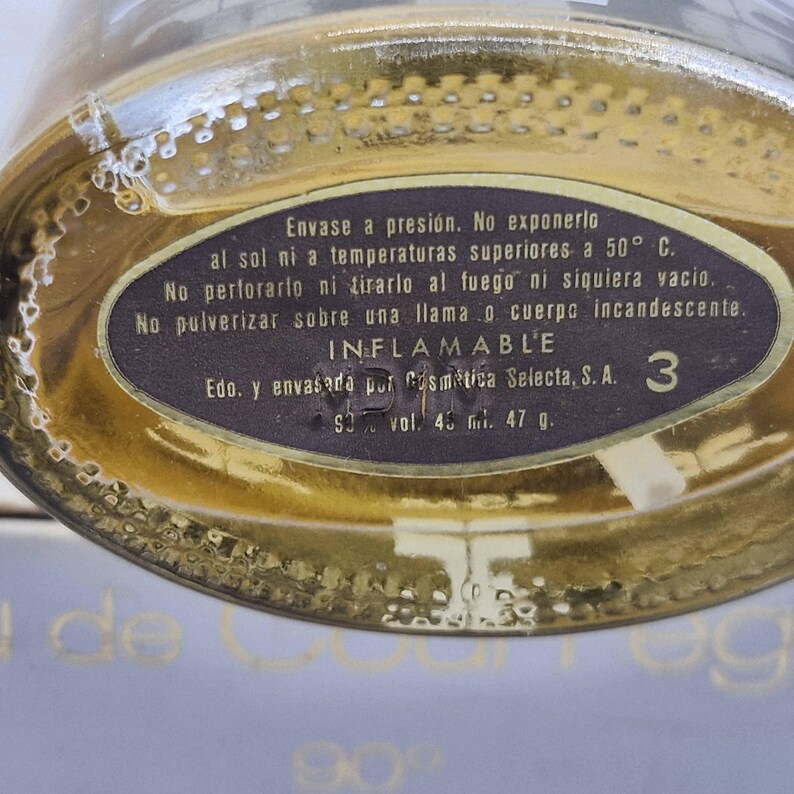 May include: Close-up of a perfume bottle with a gold-colored liquid. The bottle has a dark label with Spanish text, including warnings about pressure and flammability. The label also indicates a volume of 45 ml and a weight of 47 g.