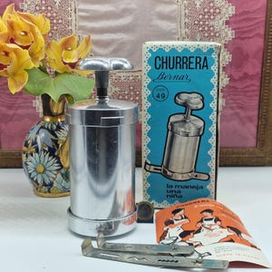 Pode incluir: Uma churrera prateada vintage, uma ferramenta para fazer churros, com uma caixa que diz "CHURRERA Bernar". A caixa é azul clara com borda branca. Um pequeno livreto de instruções e um cortador de churros também são visíveis.