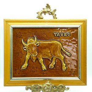 May include: A framed ceramic tile with a brown bull relief sculpture and the word "TAURO" in gold lettering. The tile is on a gold metal stand.