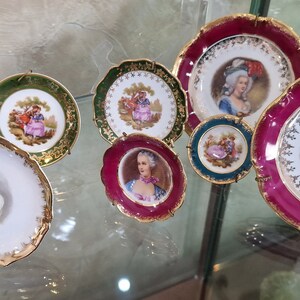 May include: A collection of seven decorative porcelain plates with gold trim. Each plate features a different portrait of a person in period clothing. The plates are displayed on a glass shelf.