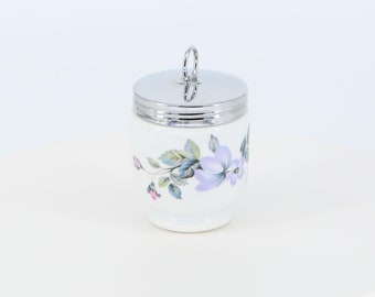 May include: A white ceramic egg cup with a floral design and a silver lid with a handle. The floral design features purple and pink flowers with green leaves.