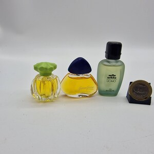 Puede incluir: Una colección de frascos de perfume en miniatura con tapas y líquidos de colores. Los frascos tienen varias formas y tamaños, uno de ellos etiquetado como "milla schon UOMO". Una moneda descansa sobre un pequeño soporte negro.