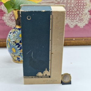 May include: A vintage box with a blue and beige design. The blue section features a stylized illustration of a building and a sun. The text "maderas de oriente" and "MYFURGIA ESPAÑA" are visible. A coin rests in front of the box.