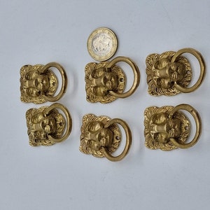 Peut inclure: Six tirettes de tiroir en forme de tête de lion dorées avec des poignées en anneau. Les tirettes sont ornées et détaillées avec un visage de lion au centre et une poignée en anneau sur le côté.
