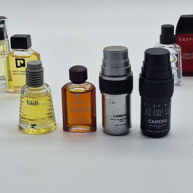 May include: A collection of miniature perfume bottles. The bottles vary in shape, color, and design, with some featuring black, silver, and brown caps. One bottle is labeled "NIAGARA", and two others are labeled "CAMERA". The bottles are arranged on a white surface.