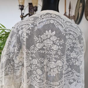 May include: Cream-coloured lace shawl with detailed floral embroidery. The delicate fabric features intricate patterns and a scalloped edge. The shawl is draped over a mannequin, highlighting its design and texture. A vintage-inspired garment.