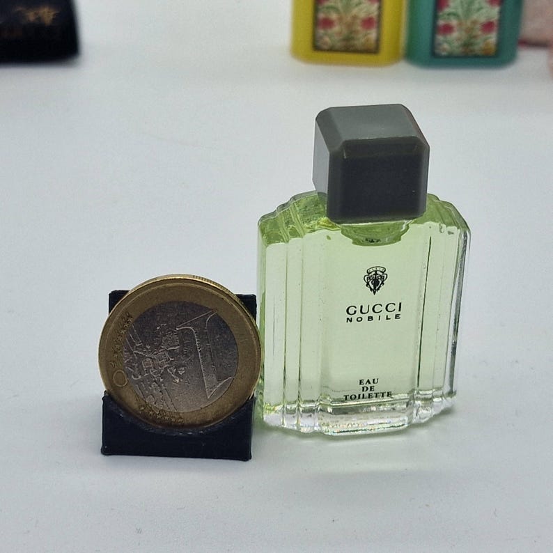 May include: A miniature Gucci Nobile Eau de Toilette bottle with a dark gray cap. The bottle is light green and has the Gucci logo and text. A one Euro coin is next to the bottle, in a black holder.