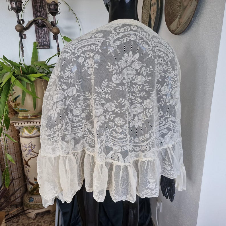 May include: An off-white, vintage lace shawl with floral embroidery and a ruffled hem. The delicate fabric is draped over a black form, showcasing the intricate details of the design. The shawl's sheer quality allows the background to be partially visible.