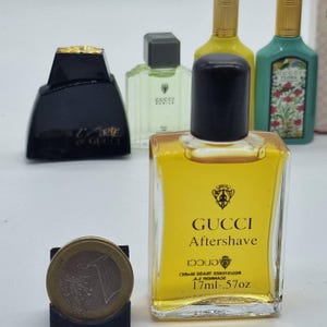 May include: A collection of Gucci miniature perfume bottles, including a clear bottle of Gucci Aftershave, a black bottle with gold accents, and other bottles with green and yellow liquid. A one Euro coin is in the foreground.