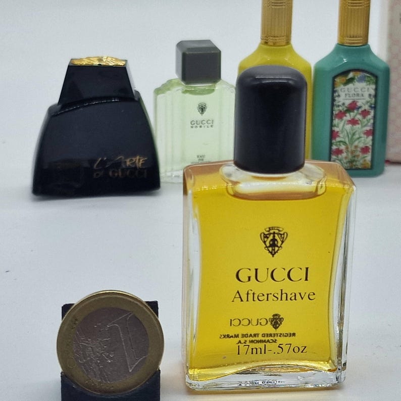 May include: A collection of Gucci miniature perfume bottles, including a clear bottle of Gucci Aftershave, a black bottle with gold accents, and other bottles with green and yellow liquid. A one Euro coin is in the foreground.